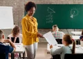 Professora entrega atividade para aluna em sala de aula durante avaliação, representando os desafios e a realidade da profissão docente.