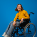 Jovem sorridente em cadeira de rodas com blusa amarela em fundo azul.
