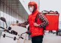 Entregador de capacete e colete vermelhos ajusta a mochila térmica ao lado de uma moto na rua