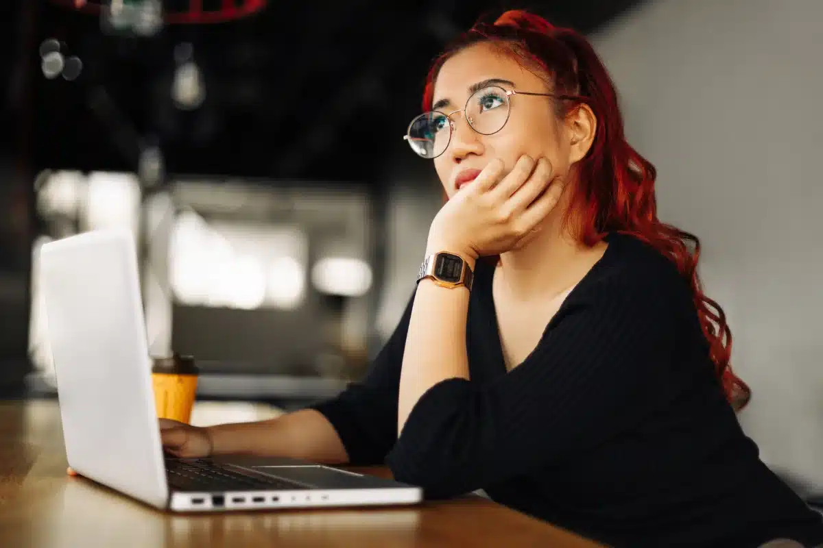 Mulher com cabelo vermelho pensativa, apoiando o rosto enquanto olha para o computador