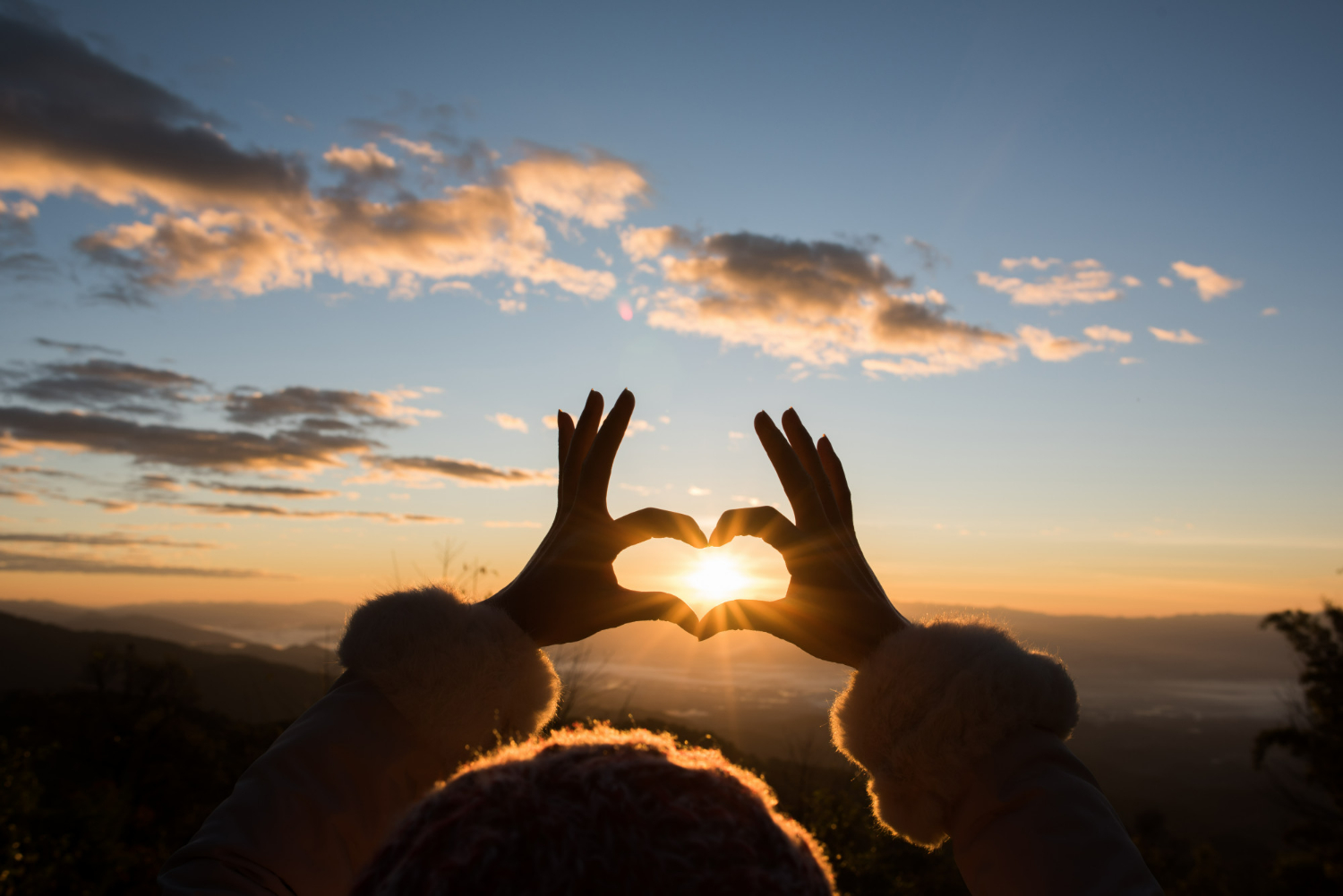 Mãos formando um coração emoldurando o sol ao pôr do sol, com uma paisagem montanhosa ao fundo