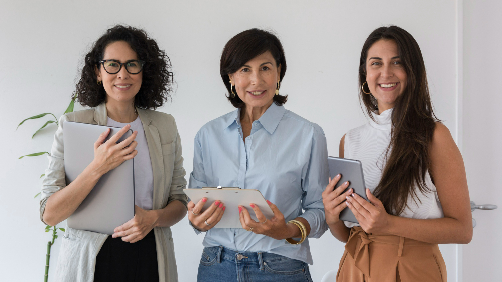 Três mulheres profissionais sorridentes segurando dispositivos eletrônicos, representando liderança e empoderamento feminino no ambiente de trabalho