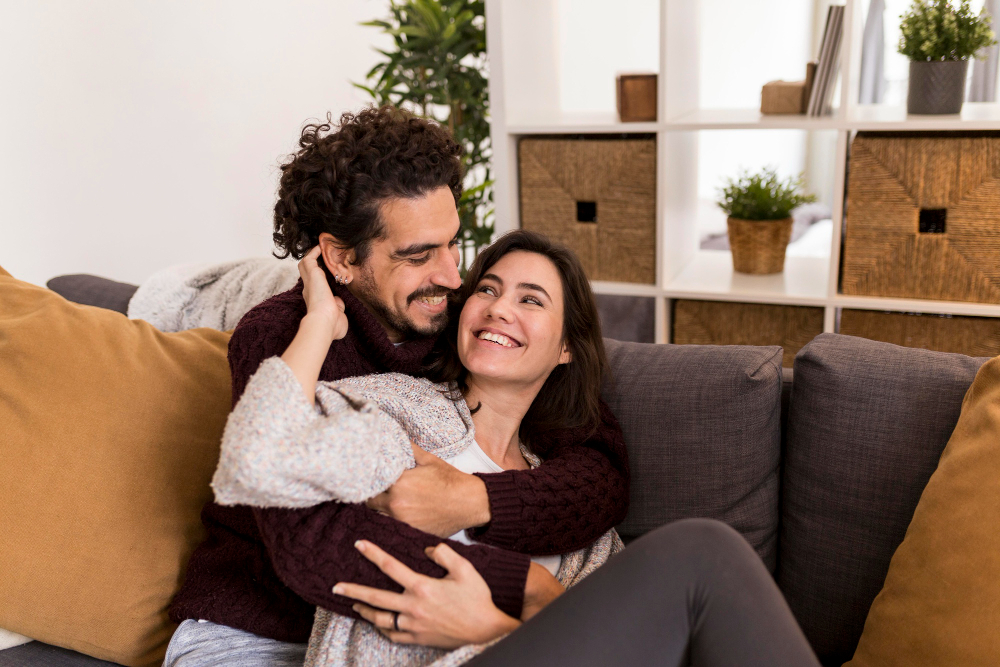 Jovem casal feliz sentado na sala de casa abraçados e sorrindo