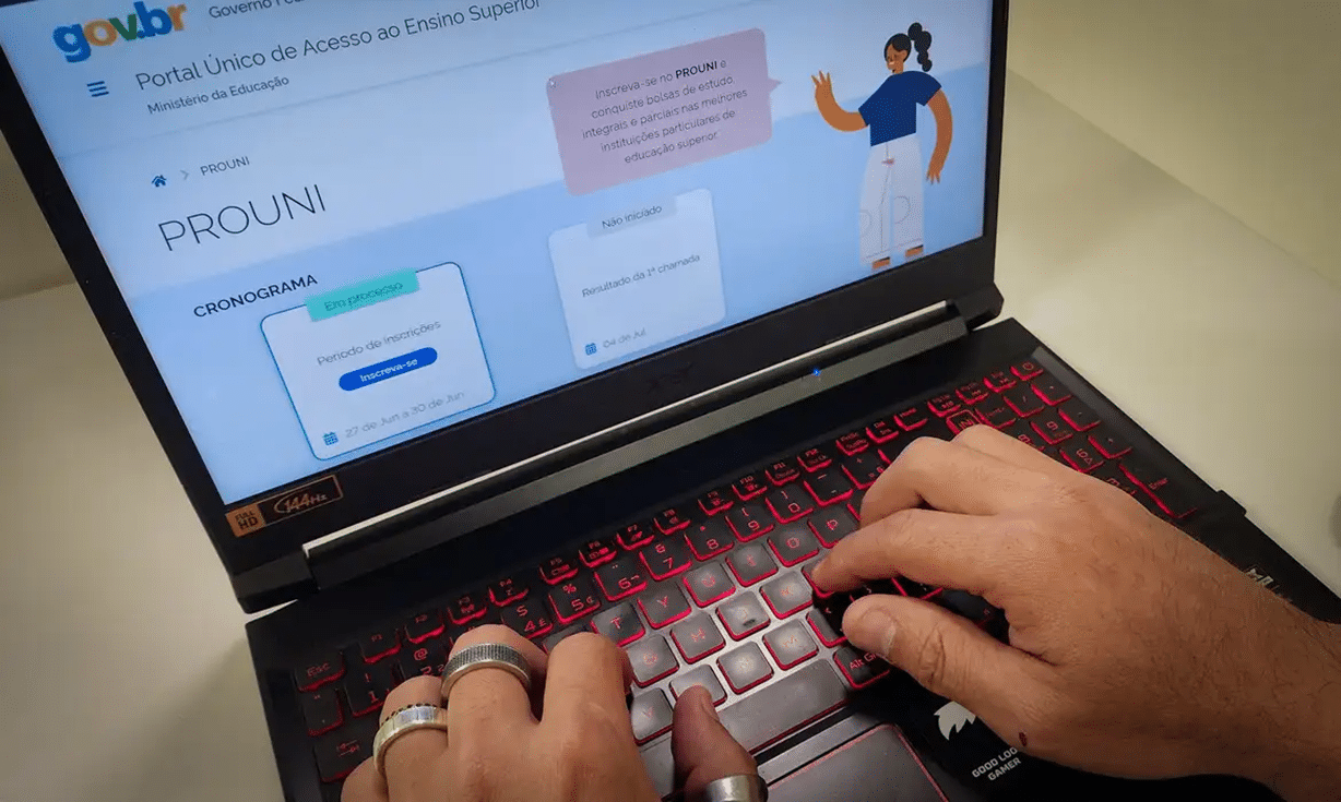 Pessoa acessando o site do Prouni em um notebook com teclado retroiluminado vermelho. A tela exibe informações sobre o período de inscrições para bolsas de estudo no ensino superior