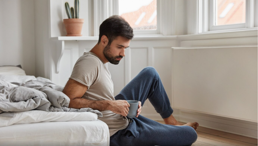 Homem sentado no chão, tomando café e refletindo, simbolizando o impacto das condições de vida na satisfação pessoal.