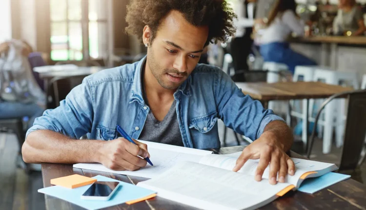 Jovem estudando com livros e anotações em um ambiente de café, preparando-se para um processo seletivo.na leitura de um livro e fazendo anotações