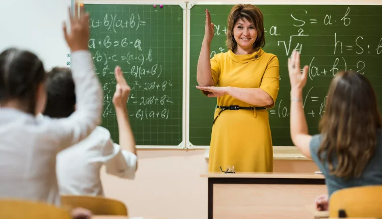 Professora sorridente interagindo com alunos em sala de aula, com quadro negro ao fundo repleto de equações matemáticas.