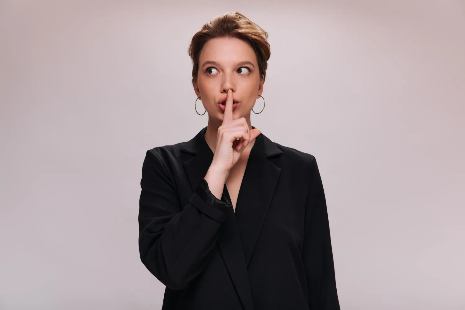 Mulher em traje preto fazendo o gesto de silêncio com a mão sobre os lábios.