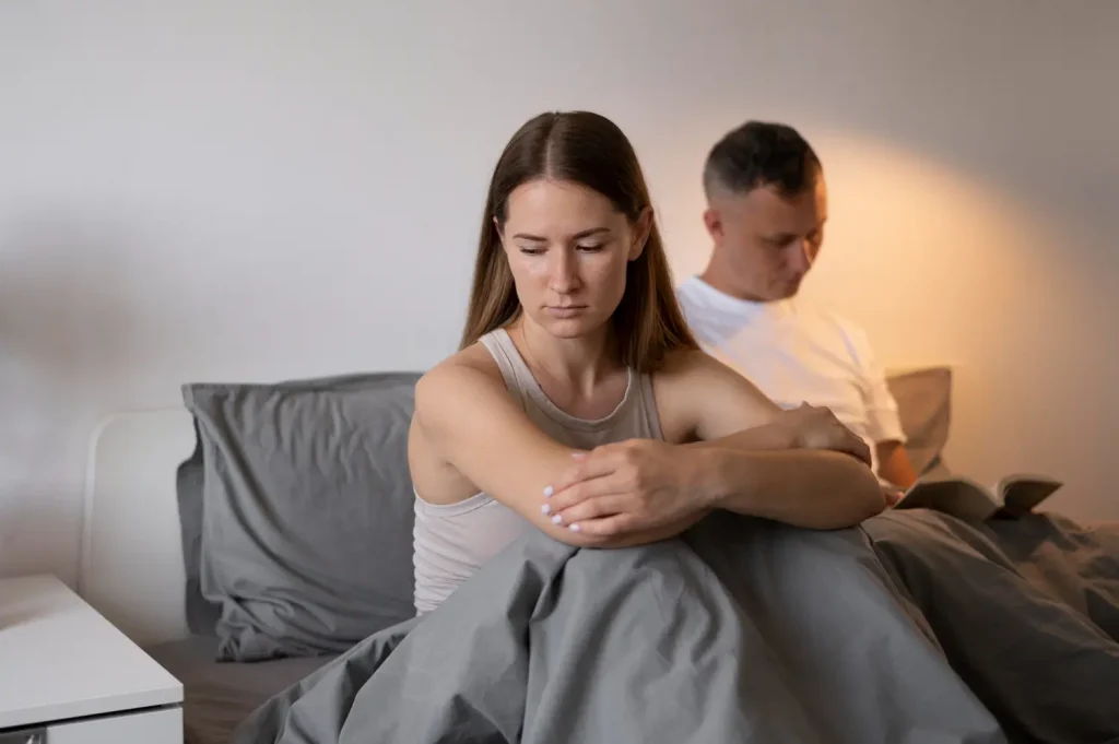 Mulher com expressão preocupada, abraçando o próprio corpo, enquanto o parceiro está ao fundo, focado em um livro, refletindo uma possível tensão emocional no relacionamento.