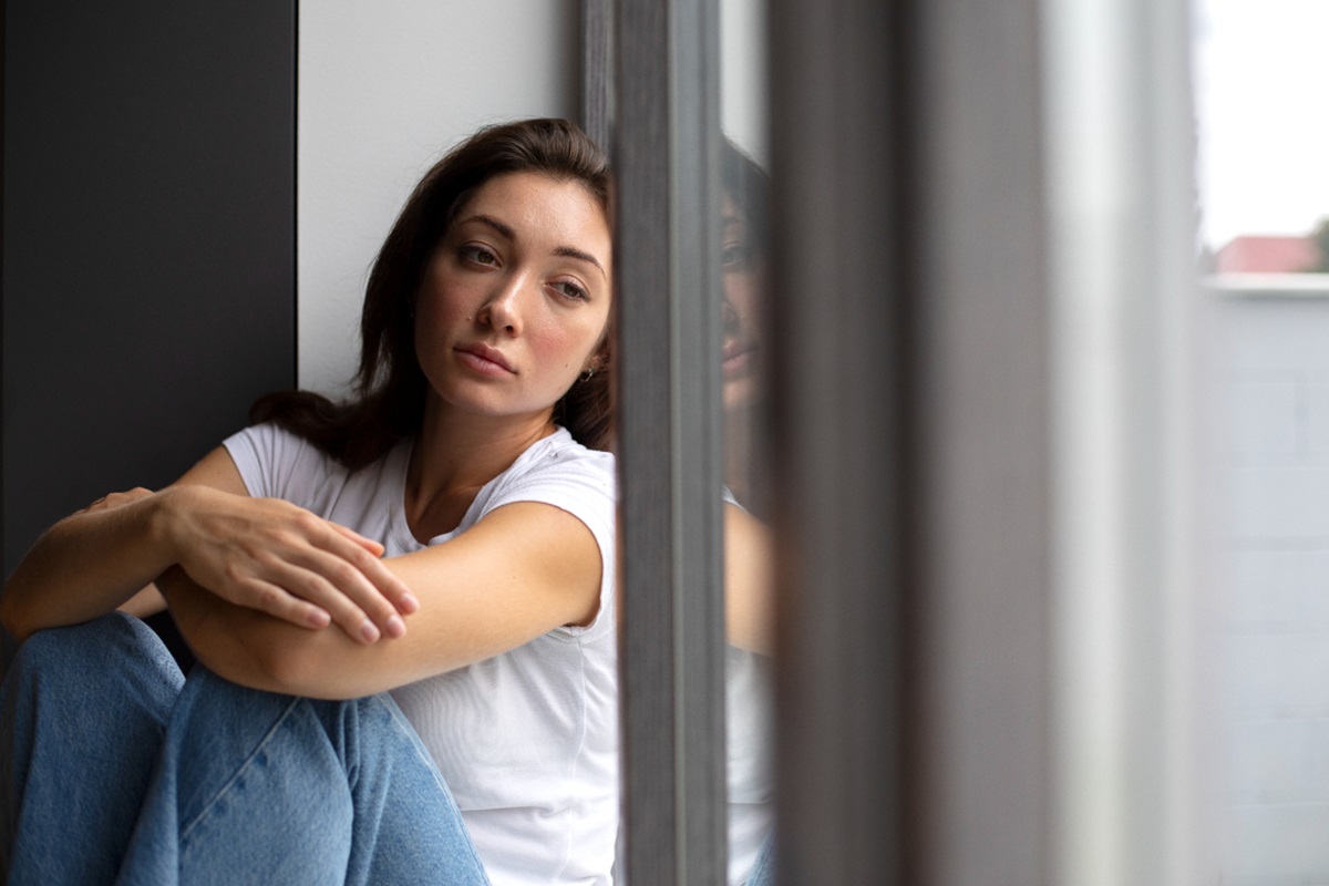 Mulher sozinha, sentada perto de uma janela, refletindo sobre a solidão e o impacto da falta de amizades.