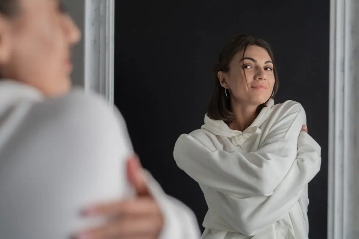 A imagem mostra uma mulher sorrindo enquanto se olha no espelho, refletindo a importância de se aceitar e valorizar. Este momento simboliza como palavras positivas podem melhorar a percepção de si mesma e aumentar o amor próprio.