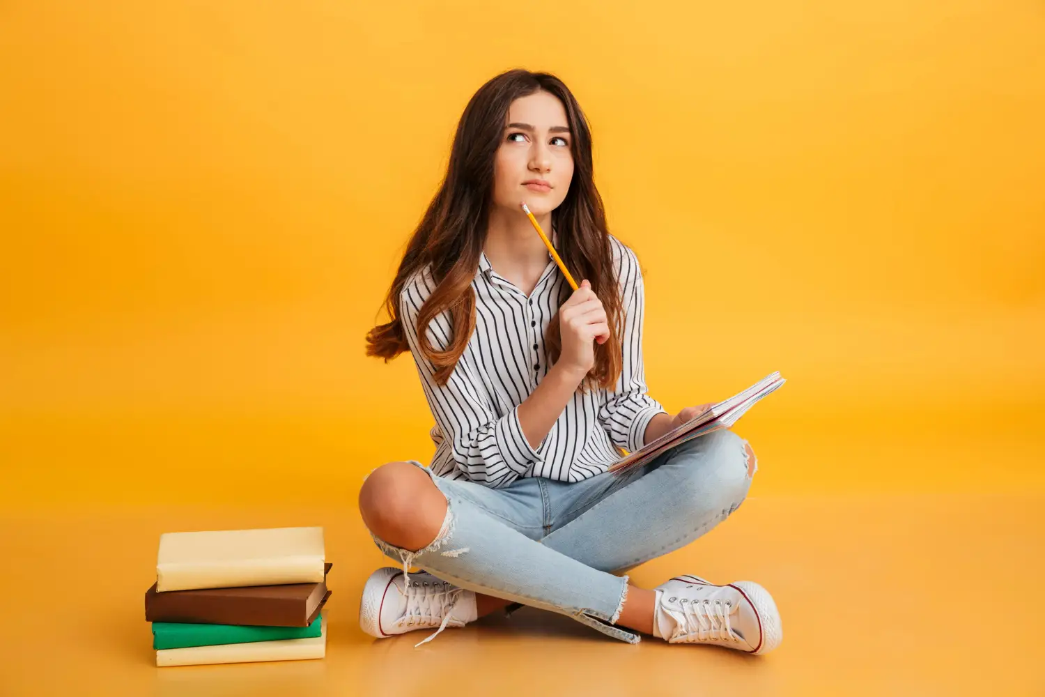 Jovem pensativa, sentada no chão com livros ao lado, refletindo sobre a formação do plural de substantivos compostos.