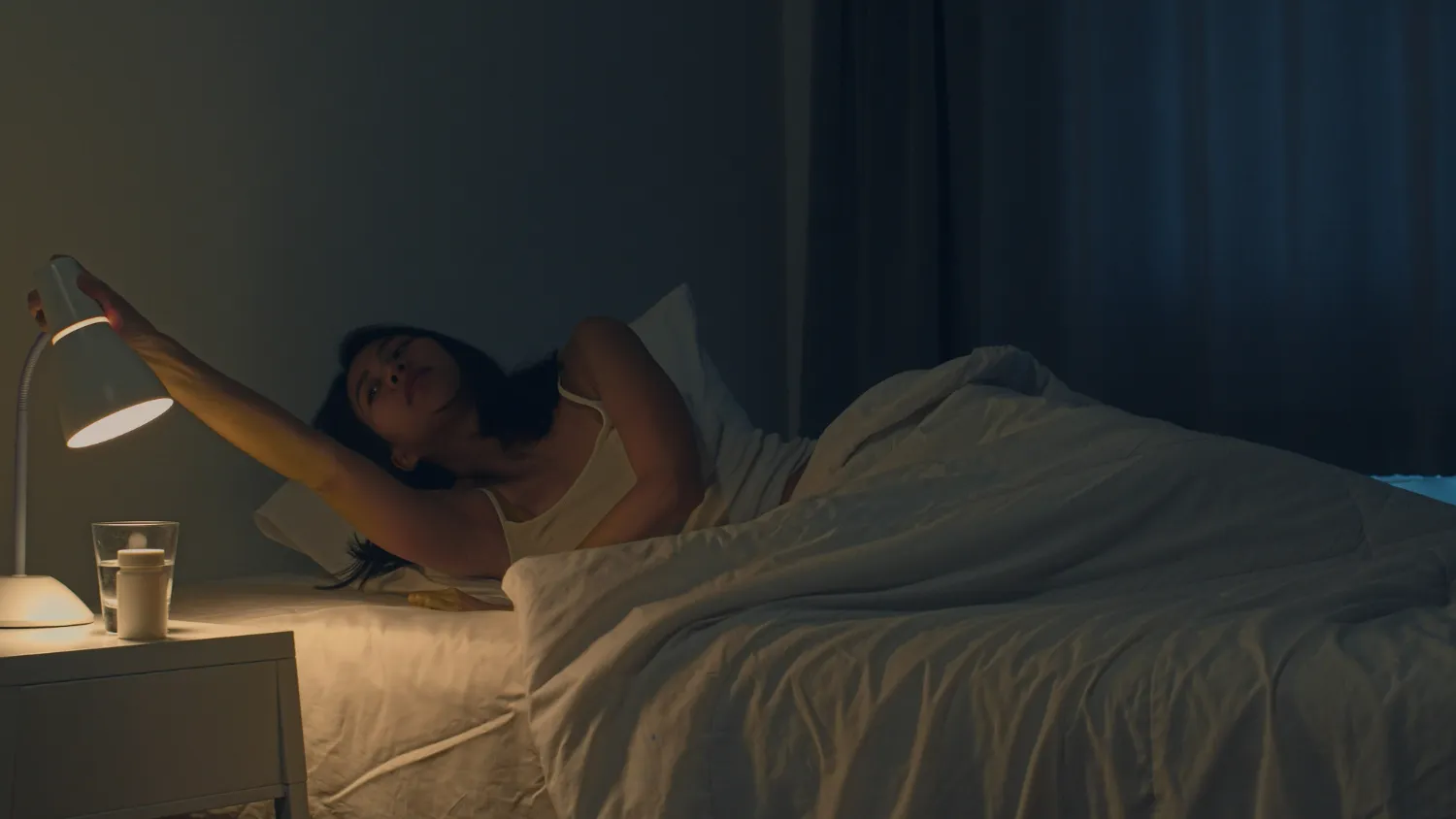 Pessoa acordando e ajustando a lâmpada ao lado da cama em um quarto escuro.