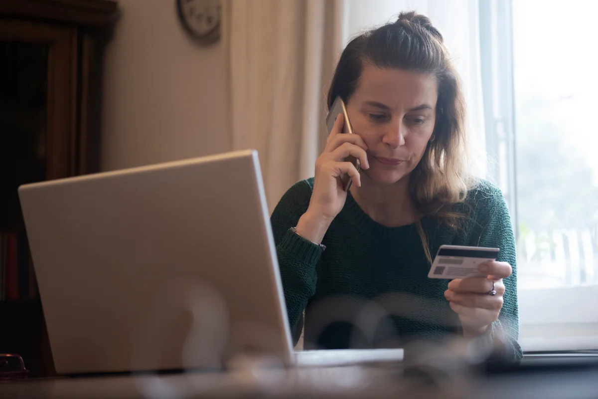 Mulher conversando ao telefone enquanto segura um cartão de crédito e usa um computador portátil.