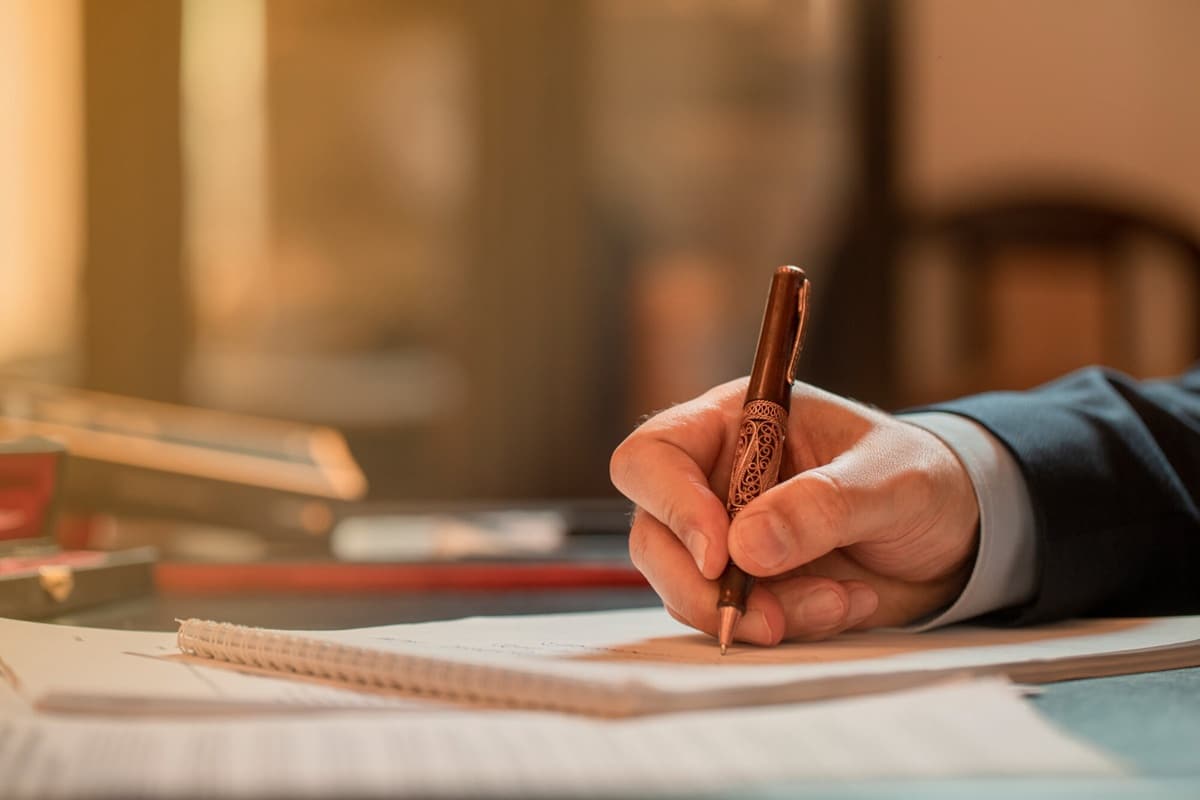 Mão escrevendo com caneta em caderno, ambiente com luz quente e foco na escrita