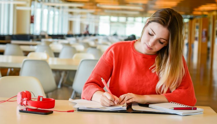 Jovem estudante concentrada escrevendo em caderno em biblioteca ampla, representando preparação para concursos públicos de nível médio.