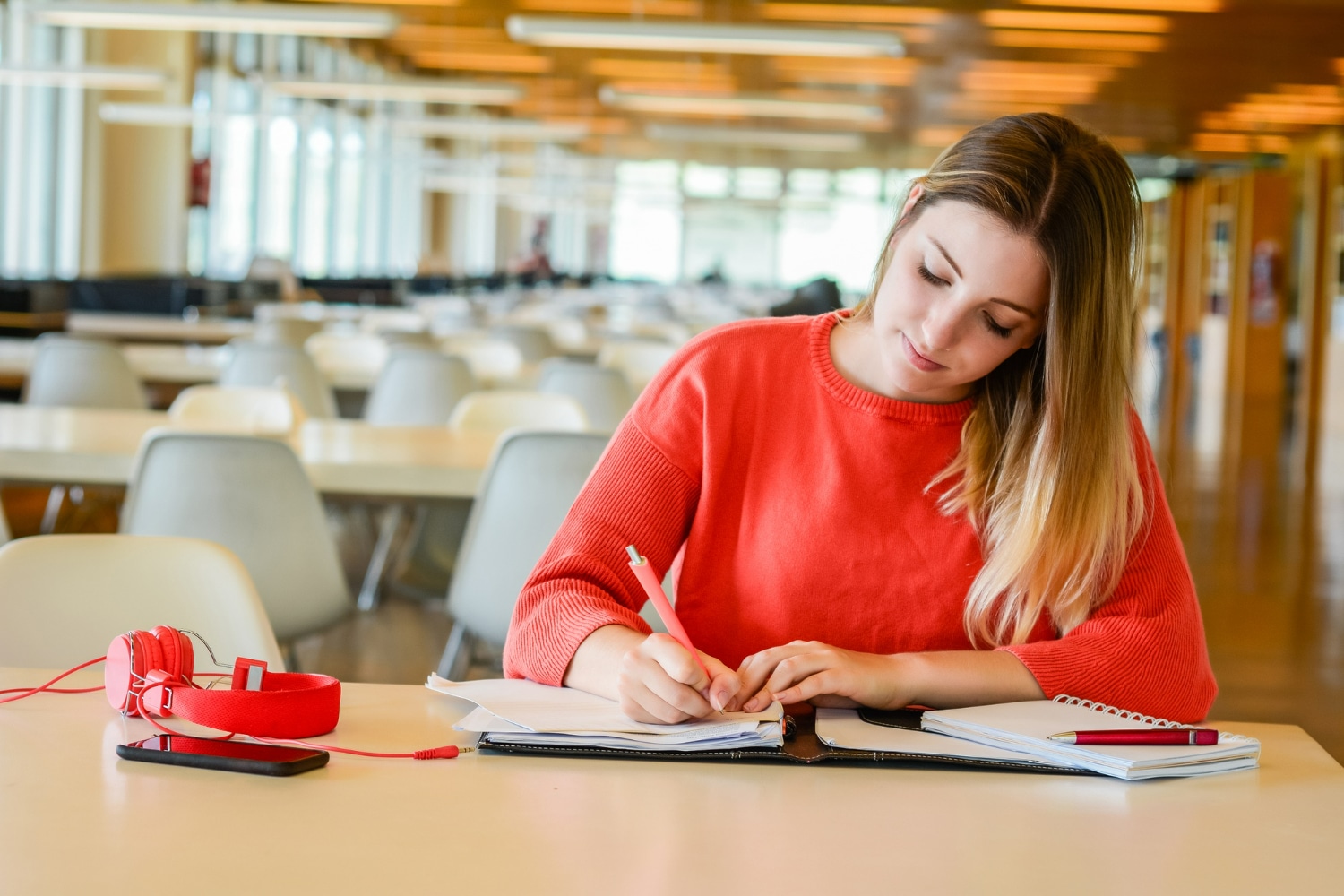 Jovem estudante concentrada escrevendo em caderno em biblioteca ampla, representando preparação para concursos públicos de nível médio.