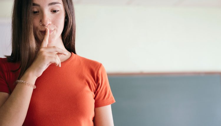 Jovem mulher com camiseta laranja fazendo sinal de silêncio com o dedo indicador nos lábios.