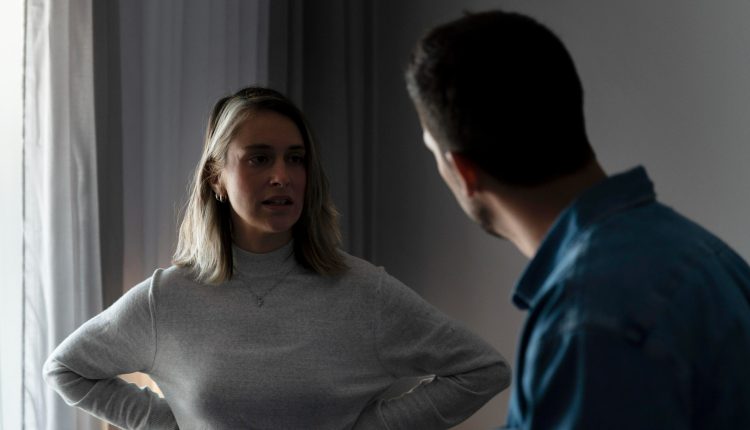 Mulher com expressão séria conversando com homem, demonstrando linguagem corporal de defesa com mãos na cintura.