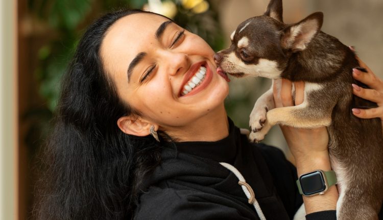 Mulher jovem segurando um cachorro pequeno que lambe seu rosto, ambos demonstrando afeto.