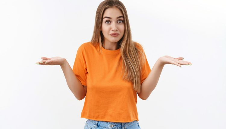 Mulher jovem com expressão de dúvida, vestindo camiseta laranja e jeans, com as mãos abertas como se não soubesse a resposta.