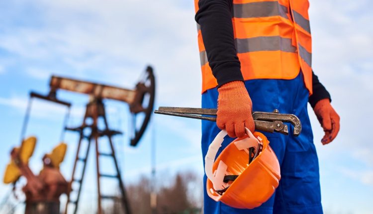Trabalhador da Petrobras segurando capacete e ferramenta com plataforma de petróleo ao fundo