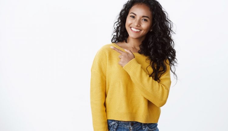 Jovem sorridente com cabelo cacheado e suéter amarelo, apontando para lado vazio