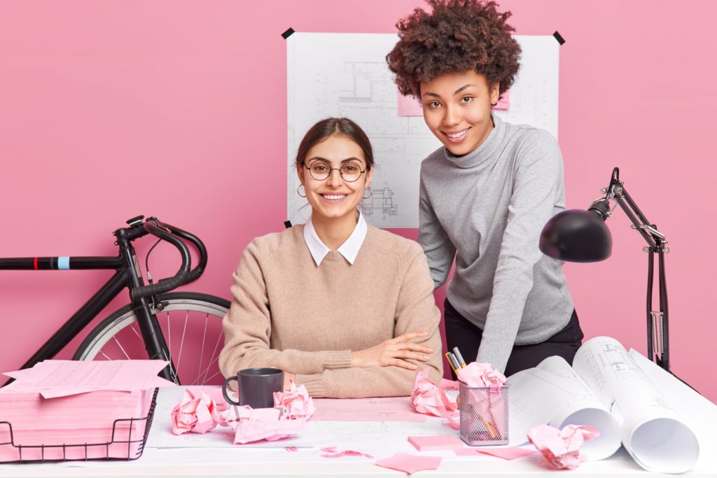 Duas mulheres sorrindo enquanto trabalham juntas em projetos de empreendedorismo, com planos e anotações em papel rosa sobre a mesa