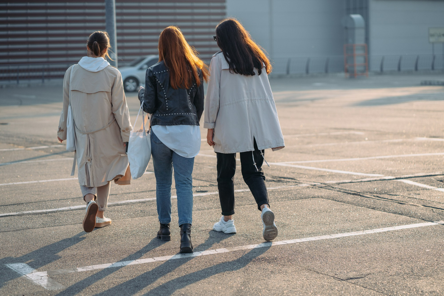 Três mulheres caminhando lado a lado em um estacionamento vazio, vistas de costas, com luz suave do pôr do sol.