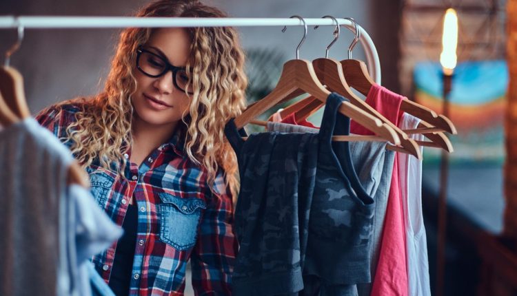 Jovem de cabelos loiro e blusa quadriculada, atrás de uma arara de cabide de roupas escolhendo uma peça.