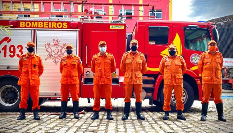 Seis bombeiros uniformizados em roupa laranja, usando máscaras, em frente a um caminhão de bombeiros vermelho.