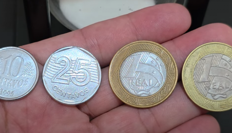 Mão segurando quatro moedas brasileiras: 10 centavos de 1995, 25 centavos, 1 real de 2012 e 1 real de 1998.