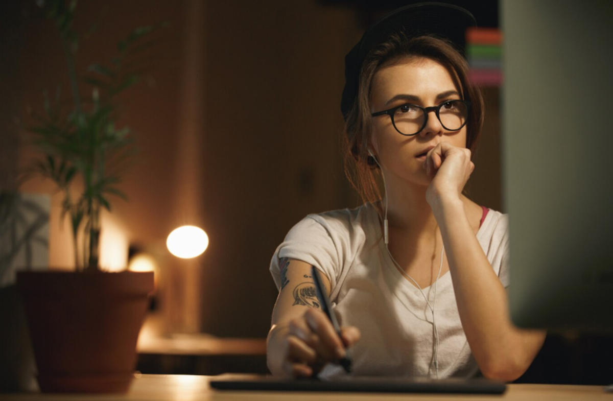 Mulher jovem com óculos, estudando e pensando em frente ao computador à noite