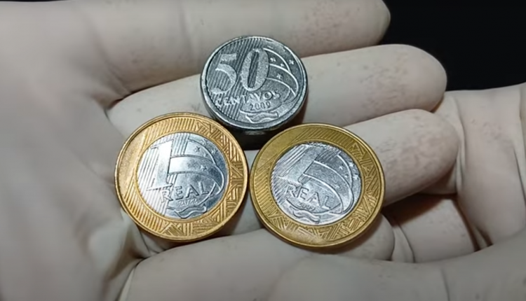 Moedas brasileiras com defeitos, incluindo 1 real, 5 reais e 50 centavos em mãos com luvas