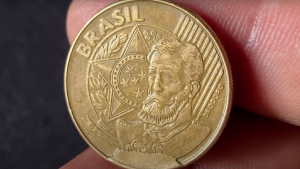 Moeda brasileira de 25 centavos com cunho quebrado, destacada em dedos, indicando seu valor colecionável potencial.