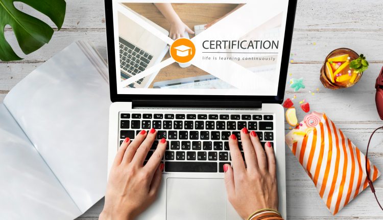 Pessoa digitando em notebook com tela mostrando palavra "CERTIFICATION" e símbolo de graduação, ao lado itens de estudo e lanche