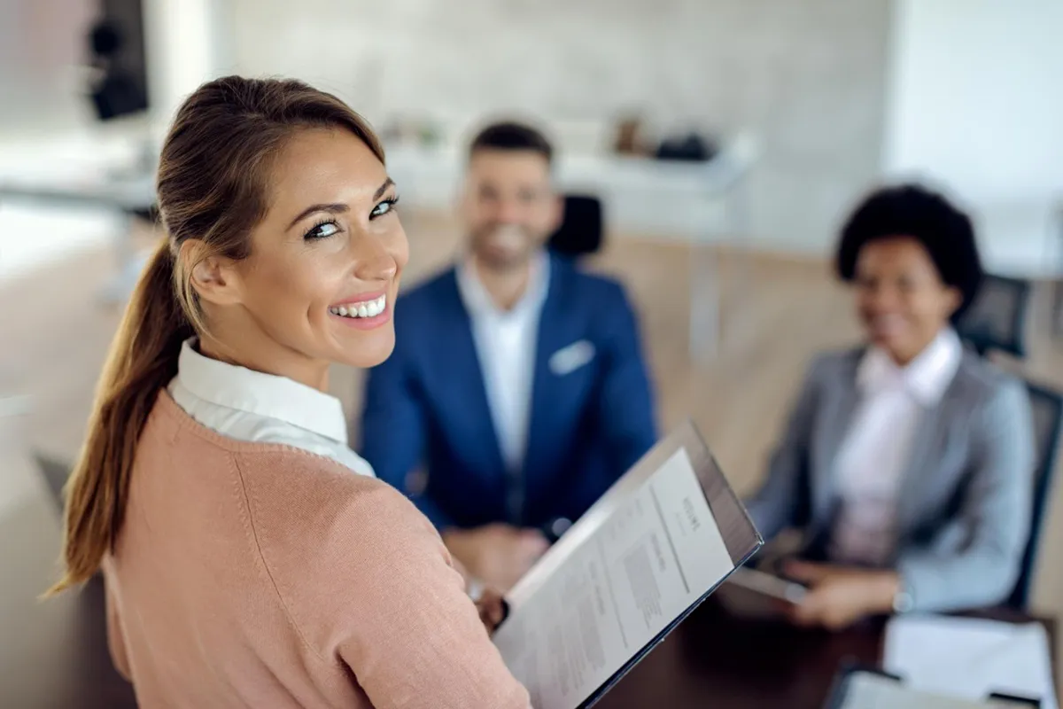 Mulher sorrindo segurando seu currículo em frente a uma mesa com outros profissionais durante uma feira de empregos.