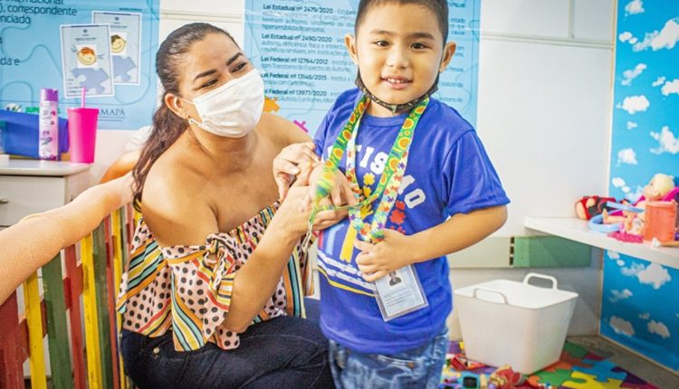 Menino usando cordão de girassol com identificação, acompanhado por mulher com máscara em ambiente de brinquedos.