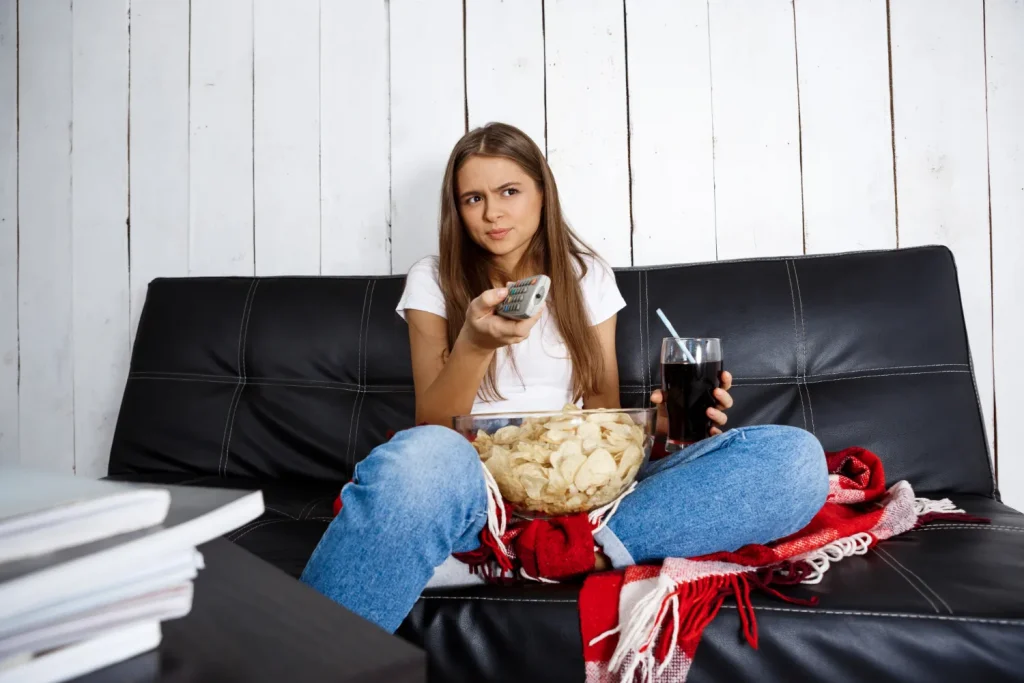 Jovem assistindo TV com expressão pensativa, segurando um copo de refrigerante e uma tigela de batatas chips