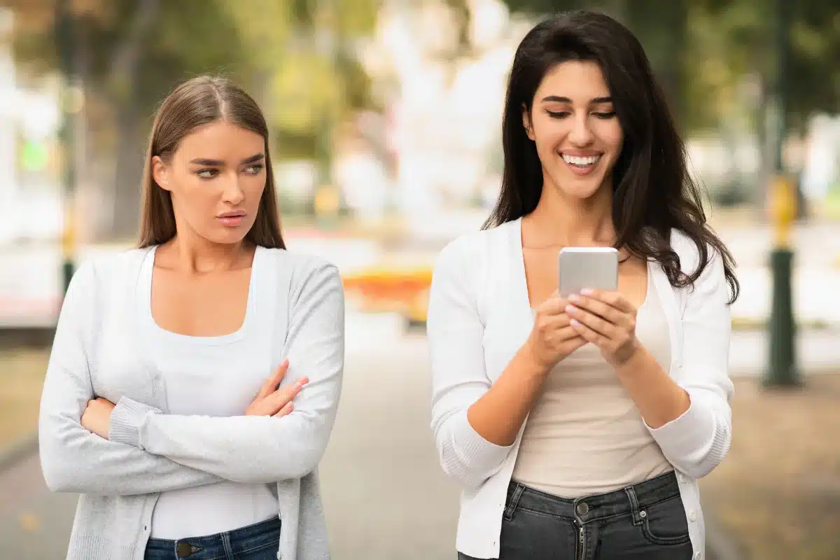 Amigas em uma caminhada, com uma parecendo desconfortável e a outra sorrindo com o celular.