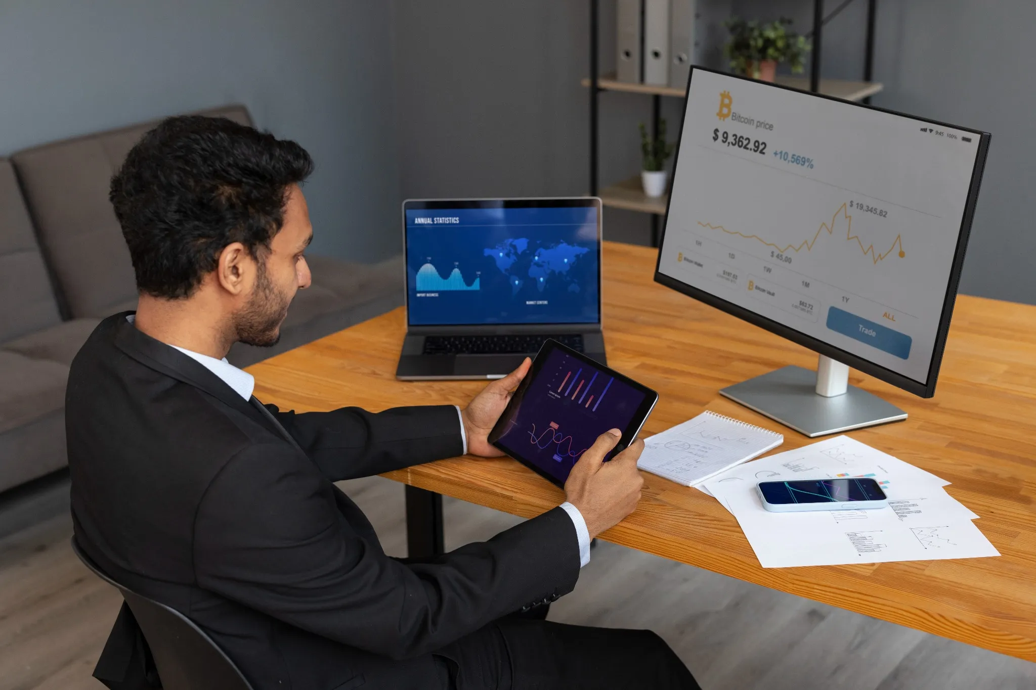 Profissional analisando gráficos financeiros e o preço do Bitcoin em uma tela de computador e tablet.