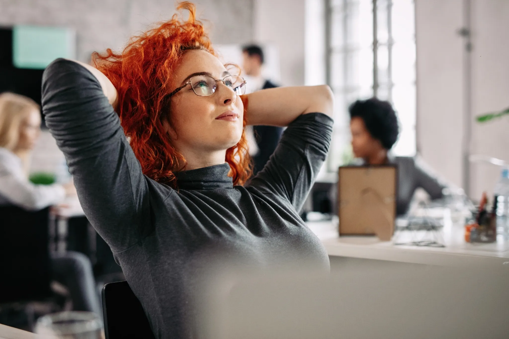 Mulher com cabelo ruivo relaxando no escritório, com expressão tranquila e pensativa enquanto descansa após o trabalho.