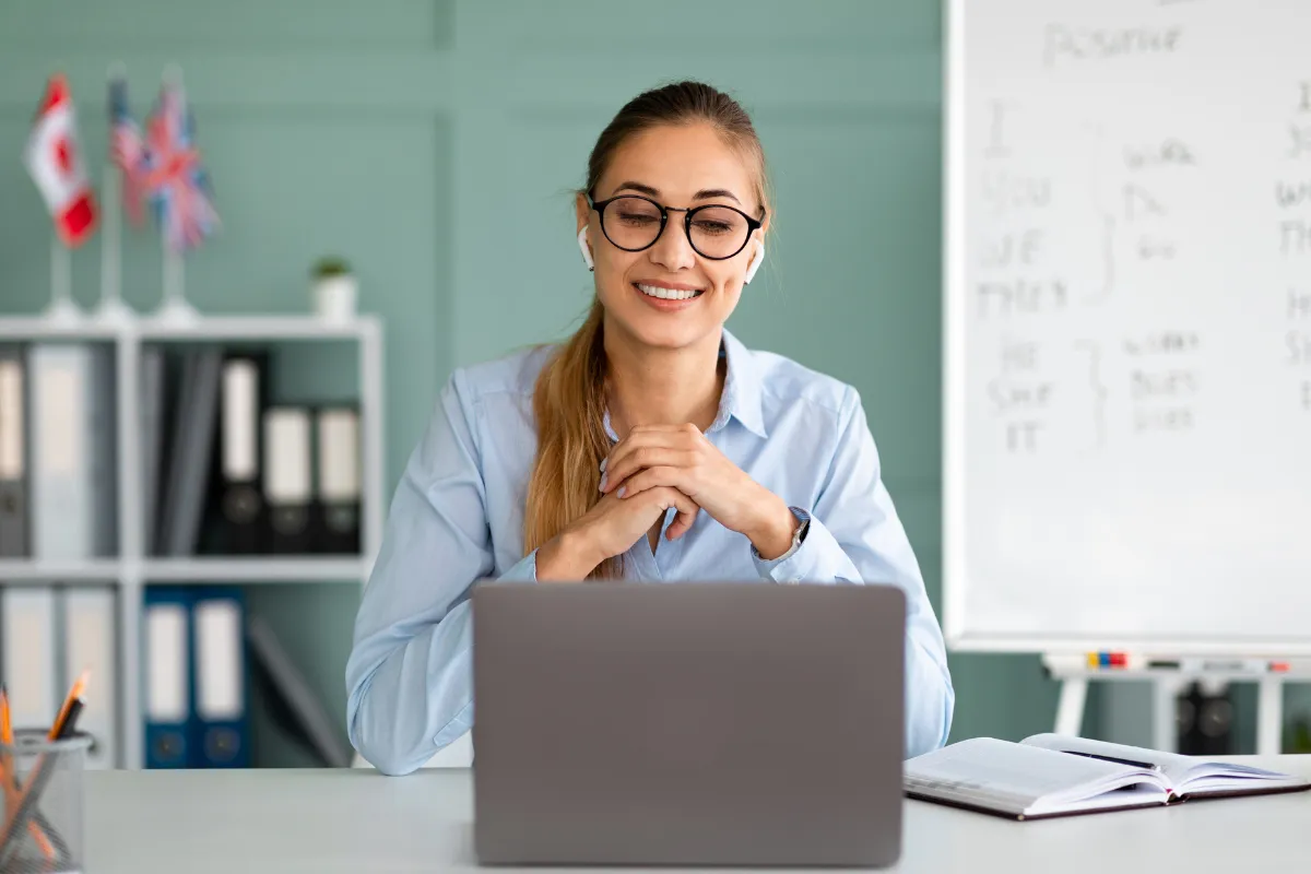 Mulher sorrindo, usando óculos e fones de ouvido, em frente a um laptop em uma sala de aula moderna.