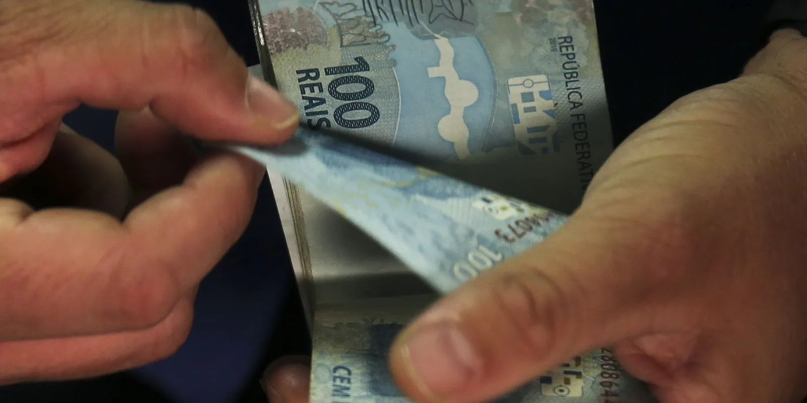 Nota de 100 reais sendo manuseada por mãos, representando transação financeira.
