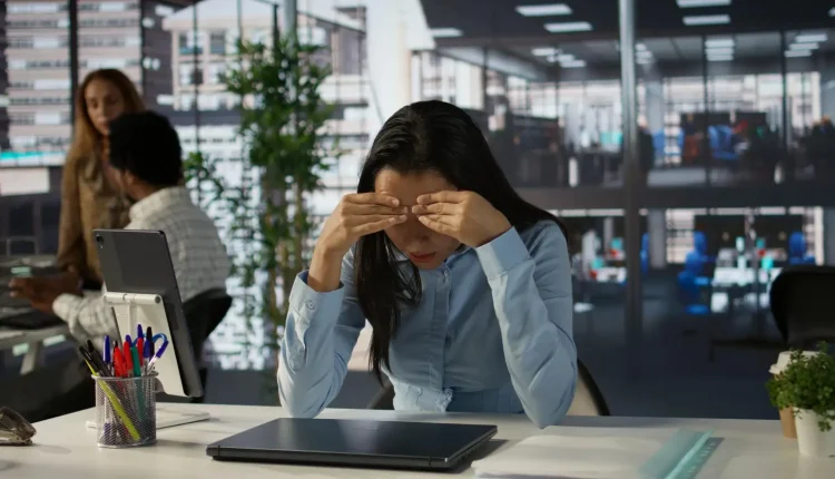 Mulher no escritório com sinais de Burnout, segurando a testa, visivelmente estressada e exausta