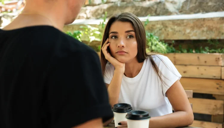 Mulher demonstrando desinteresse em uma conversa enquanto segura um copo de café, olhando para o lado enquanto o homem, em pé, fala com ela.
