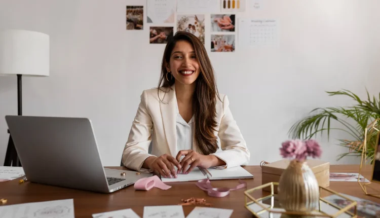 Mulher sorridente em escritório moderno, usando blazer branco, com laptop e materiais de trabalho à frente