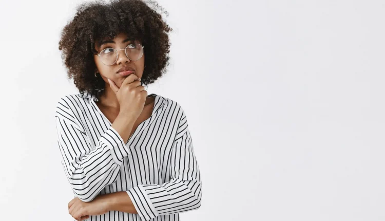 Mulher pensativa com cabelo cacheado, usando óculos e fazendo gesto de reflexão, com o rosto voltado para a direita.