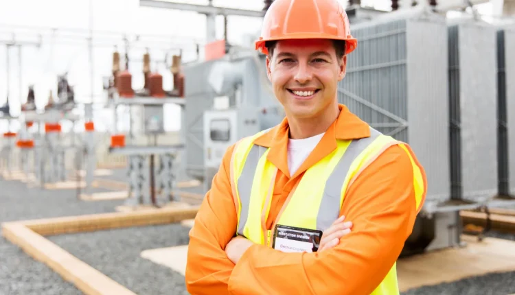 Engenheiro de segurança em campo, com uniforme de segurança e capacete, sorrindo em frente à estação de energia.