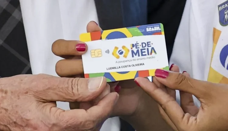 Cartão do programa Pé-de-Meia sendo entregue por duas pessoas, com foco nas mãos e no cartão colorido com o nome Ludmilla Costa Oliveira.